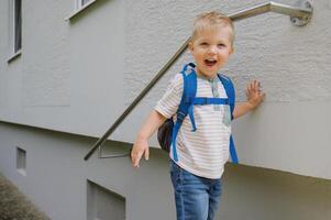 Happy cute kid boy with backpack standing near gray wall building in daylight photo