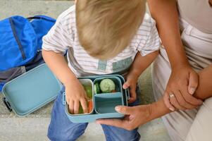 Unrecognizable mother and crop kid sitting on bench with salad lunchbox in daylight photo
