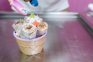 A worker is decorating a rolled ice cream tub with chips and teddy bear toppings with a spoon photo