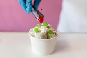 A worker is decorating a rolled ice cream tub with a cherry on top using tongs photo