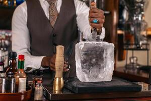 Close-up of an unrecognizable black bartender cutting a large ice cube to prepare a cocktail photo
