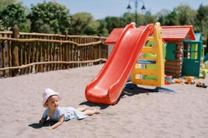 Little girl lies on the sand near a slide on the playground and looks away photo