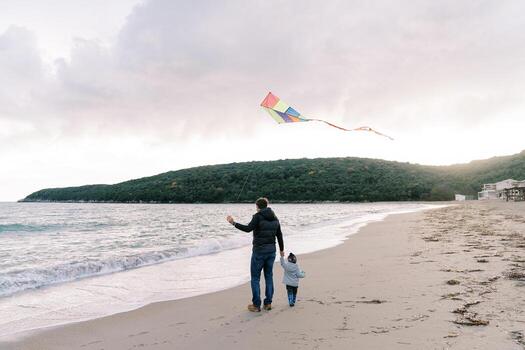 Dad with a kite on a string stands with a little girl on the seashore. Back view photo
