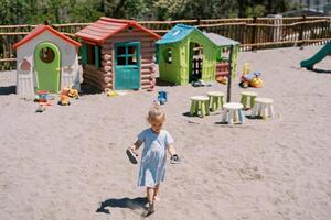 Little girl walks barefoot on the sand on the playground with sandals in her hands photo