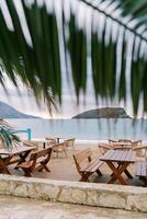 View from behind a green palm leaf on wooden tables and chairs standing on the beach by the sea photo