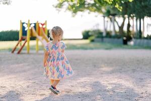 Little girl walks along the playground to a colorful slide. Back view photo