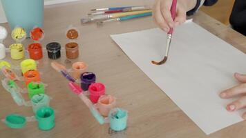 A preschool boy, 4-5 years old, takes paint with a brush and applies it to a white sheet. Sitting at the table in daylight, he enjoys experimenting with colors video