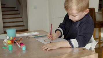 A 4-5-year-old boy dips his brush into the paint and begins drawing on a white sheet of paper. Sitting at a table in natural light, he enjoys the creative process. video