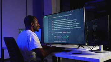 A dedicated African American software developer writes and debugs code on multiple screens in a dimly lit office video