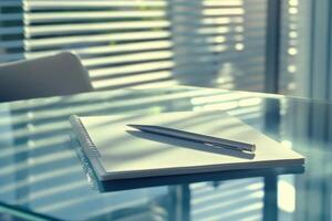 Sunlit office desk with pen and notebook on glass surface reflecting blinds photo