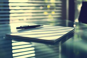 Pen and notebook on glass table with striped shadow patterns from blinds photo