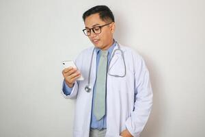 A man in a white lab coat is looking at his cell phone. He is smiling and he is enjoying himself photo