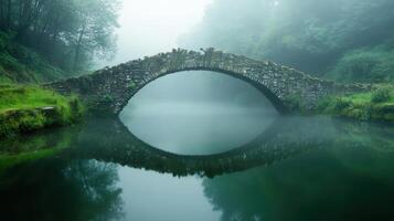 A serene stone bridge arches over a misty river, reflecting in the calm water below. photo