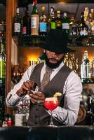 stylish black bartender in a hat serving a cocktail decorated with garnish. He is scenting the cup with a perfumer photo