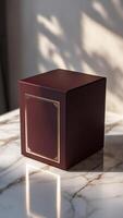 Elegant Packaging. A square dark red box with a delicate gold frame sits on a marble surface, illuminated by soft light. photo