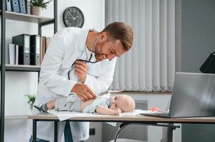 Using ,stethoscope.. Doctor in white coat is taking care of little toddler indoors photo