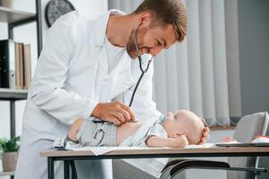 Close up view. Using stethoscope. Docto,r in white coat is taking care of little toddler indoors photo