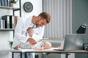 Using stethoscope. Doctor in white coat is taking care of little toddler indoors photo
