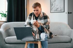 Sitting, and sneezes. Sick man is on the sofa at home by the laptop. Conception of illness photo