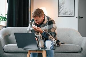 Sitting and ,sneezes. Sick man is on the sofa at home by the laptop. Conception of illness photo