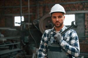 Standing in grey uniform and in white hard hat. Factory male worker is indoors photo