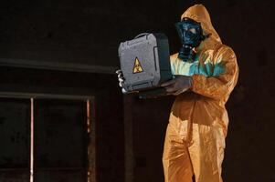 Blue light is going from ,hard case that is with hazard sign. Man dressed in chemical protection suit in the ruins of the post apocalyptic building photo