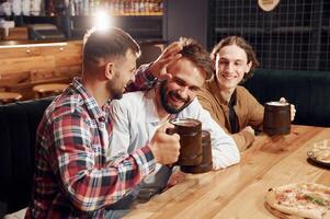 Smiling, knocking mugs, pizza on the table. Three friends are in the bar drinking beer and having a good time photo