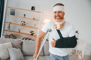 Smiling, showing thumb up. Man with crutches is at home indoors. Having injuries photo