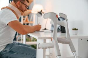 Sitting ,by ,the table and writing. Man with crutches is at home indoors. Having injuries photo