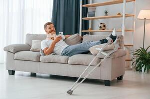 Laying down ,and resting on the sofa. Man with crutches is at home indoors. Having injuries photo