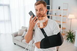 Using smartphone.. Man with crutches is at home indoors. Having injuries photo