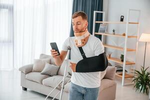 Using smartphone. Man with crutches, is at home indoors. Having injuries photo