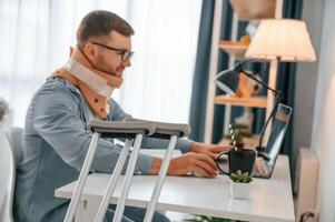 Working ,from home by using laptop. Man with crutches is indoors. Having injuries photo
