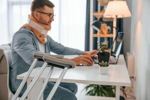 Working from home by using, laptop. Man with crutches is indoors. Having injuries photo