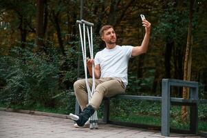 Making a selfie. Man ,with crutches is in the park outdoors. Having leg injury photo