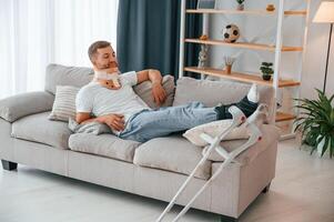 Laying down and resting on the sofa.. Man with ,crutches is at home indoors. Having injuries photo