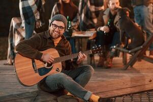Man in ,hat ,is sitting and playing guitar. Group of people is spending time together on the backyard at evening time photo