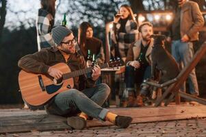 Man in hat, is sitting and playing guitar. Group of people is spending time together on the backyard at evening time photo