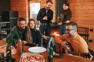Man, in glasses is playing guitar. Group of friends is having good weekend indoors in the wooden building together photo