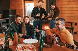 Man in glasses is playing, guitar. Group of friends is having good weekend indoors in the wooden building together photo