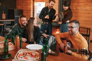 Man in ,glasses is ,playing guitar. Group of friends is having good weekend indoors in the wooden building together photo