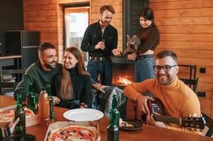 Man in ,glasses ,is playing guitar. Group of friends is having good weekend indoors in the wooden building together photo