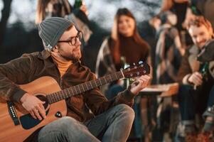 Man in hat is sitting ,and playing guitar. Group of people is spending time together on the backyard at evening time photo