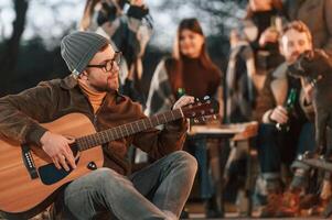 Man in hat is sitting and playing guitar. Group of people is spending time together on the backyard at evening time photo