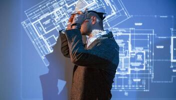 Male architect in hard ,hat ,is standing against projector with building plan on it photo