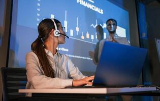 Looking at colleague. Stock broker is sitting and working by laptop. Man is standing near her, showing data on projector photo
