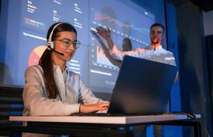 Stock broker is sitting and ,working by laptop. Man is standing near her, showing data on projector photo