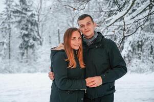 Standing and posing while embracing each other. Couple is in the winter forest, having a walk photo