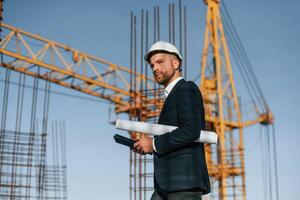 Side view,. Walking and holding paper with plan and digital tablet. Businessman in formal clothes is on the construction site photo