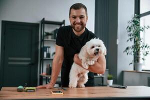 Man ,is smiling. Groomer is with Maltese dog indoors. Man is in the black uniform photo